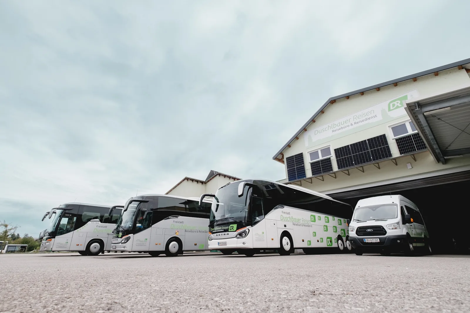 Drei Reisebusse und ein Kleinbus vor der Halle mit dem Schild Duschlbauer Reisen, Reisebüro & Reisedienst